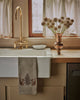 A vintage-style kitchen sink with a brass faucet, white curtain-covered window, Coral & Tusk Chipmunks with Pinecone Tea Towel, small bowl, and copper vase of dried allium flowers on a wooden countertop.