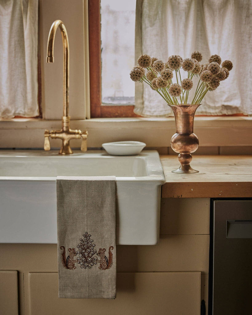 A vintage-style kitchen sink with a brass faucet, white curtain-covered window, Coral & Tusk Chipmunks with Pinecone Tea Towel, small bowl, and copper vase of dried allium flowers on a wooden countertop.