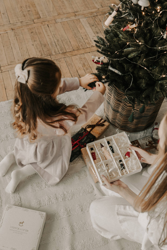 Two children sit on a white rug by a decorated Christmas tree, hanging a Sabo Concept Christmas Ornament. An open compartmentalized box with more ornaments rests nearby. The cozy scene radiates warmth and festive cheer.