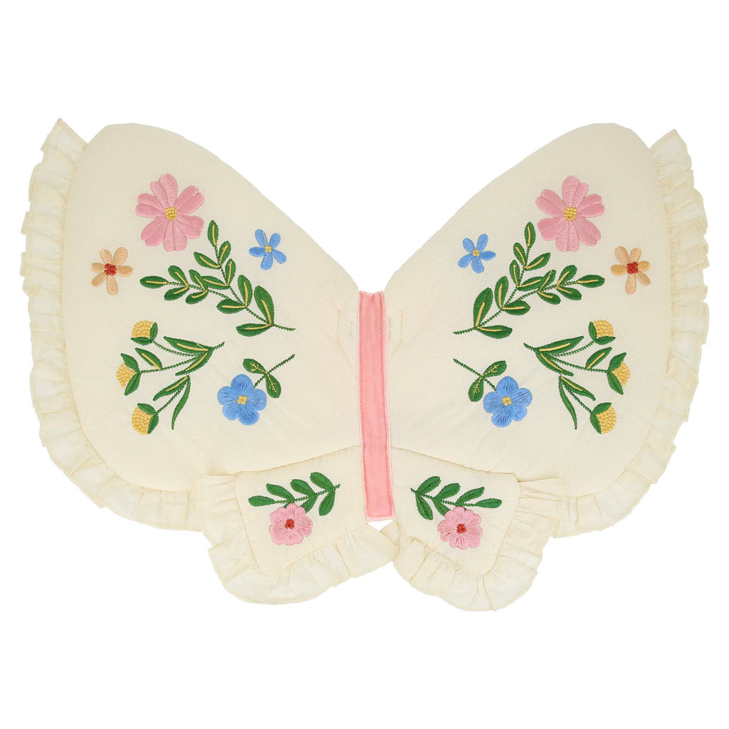 The Meri Meri Butterfly Wings pillow features a butterfly shape in cream natural cotton with ruffled edges, colorful embroidered flowers and green leaves on the wings, and a pink stripe at the center resembling a butterfly's body.