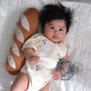 A baby with thick dark hair, in a cream onesie, rests on a blanket holding a blue toy and pacifier, lying against the plush Tummy Time Baguette pillow—ideal for tummy time and imaginative play.