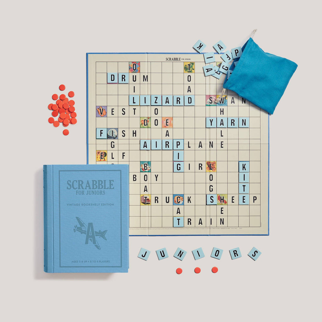 Scrabble for Juniors Vintage Bookshelf Edition featuring a blue cloth-bound storage book, a game board filled with letter tiles, a blue tile pouch, and a collection of red circular tokens.