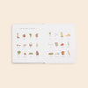 An open book titled "Our Little Adventures To the Farmers Market - Board Book" lays on a beige surface. The pages feature 24 hand-drawn illustrations of various fruits, vegetables, and other market items, each labeled with their respective names. Ideal as a baby gift, this children's book introduces early language concepts like apple, carrot, broccoli, egg, and bread.