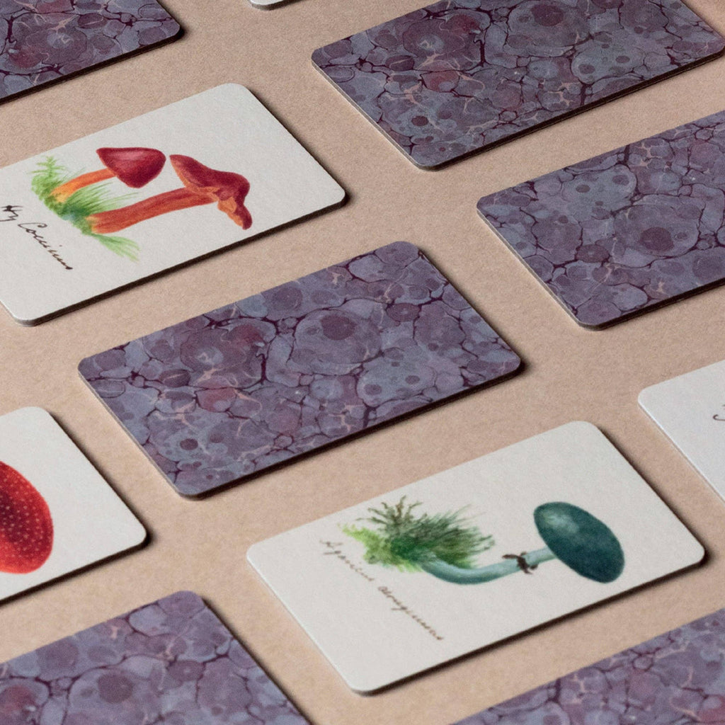 Several rectangular cards from the Memory Game - Fungi are laid on a surface, some face-down with a purple marbled pattern, others face-up showing colorful vintage fungi illustrations.