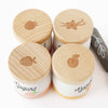 The Yogurt Play Food Pack includes four containers with wooden lids, each featuring emblems of raspberry, vanilla, strawberry, and peach. Ideal for plastic-free role play. "Yogurt" is printed on them. A black stone with a leaf design is also included.