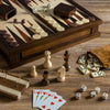 A wooden table displays assorted board game pieces, including the Heirloom Edition: 7-in-1 set (Chess, Checkers, Backgammon, Cribbage, Poker Dice, Cards, Dominoes), with all items arranged neatly in a walnut cabinet and brown pouch.