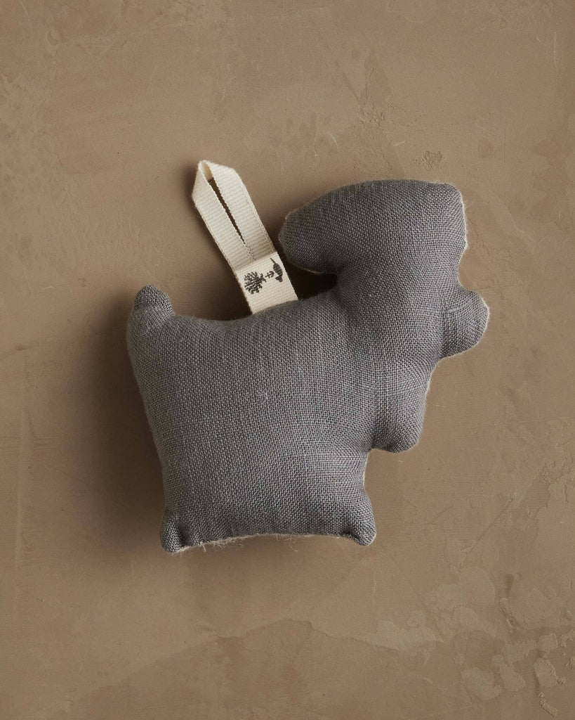 A charming Coral & Tusk Reindeer with Bells Ornament, crafted from fabric with a cream looped tag, rests on a textured light brown surface like a hidden holiday treasure waiting to be discovered.