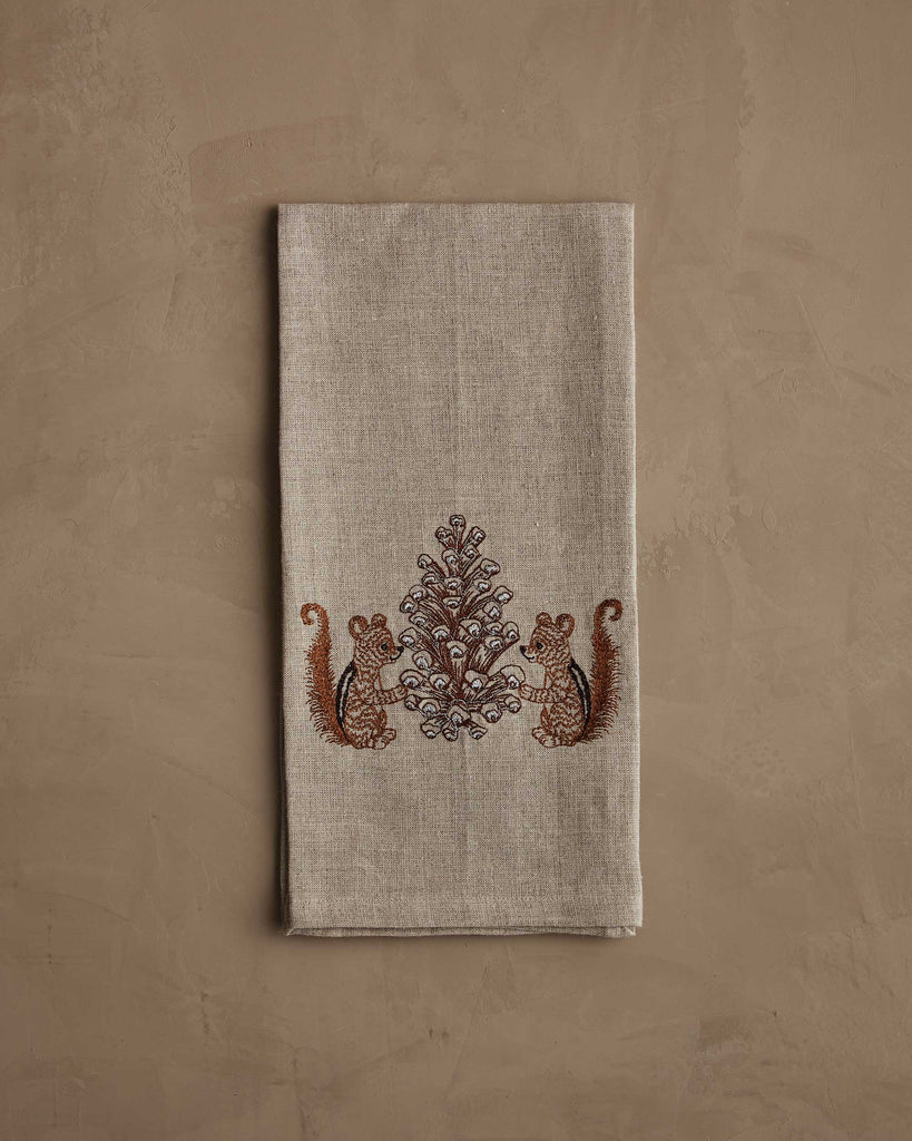 The Coral & Tusk Chipmunks with Pinecone Tea Towel features embroidered chipmunks with a pinecone and leafy motif, accented by metallic silver stitching on beige linen, displayed on a neutral textured background.