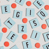 Scattered light blue square letter tiles and orange circular tokens from a Scrabble for Juniors Vintage Bookshelf Edition on a light background.