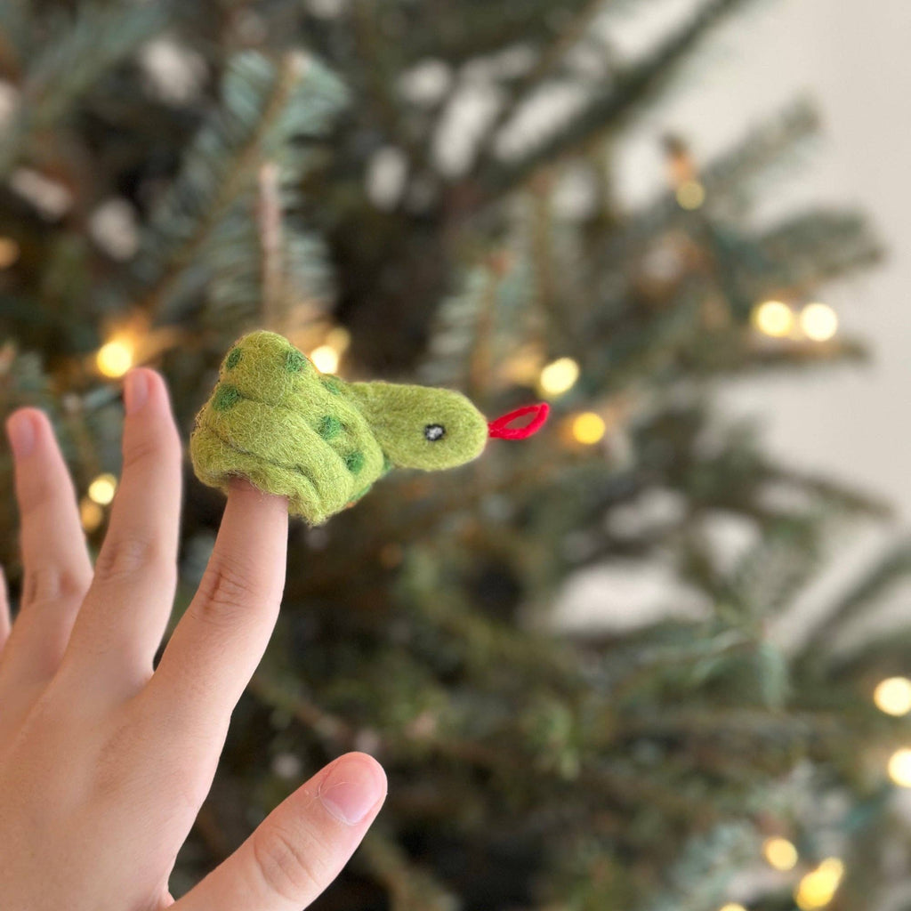 A hand wearing a Snake Finger Puppet is held up in front of a blurred, decorated Christmas tree with lights—perfect for imaginative play and creative storytelling this festive season.