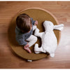 A toddler sits cross-legged on a round brown cushion beside a Senger Naturwelt Cuddly Animal - White Bunny, on a wooden floor. The photo is taken from above.