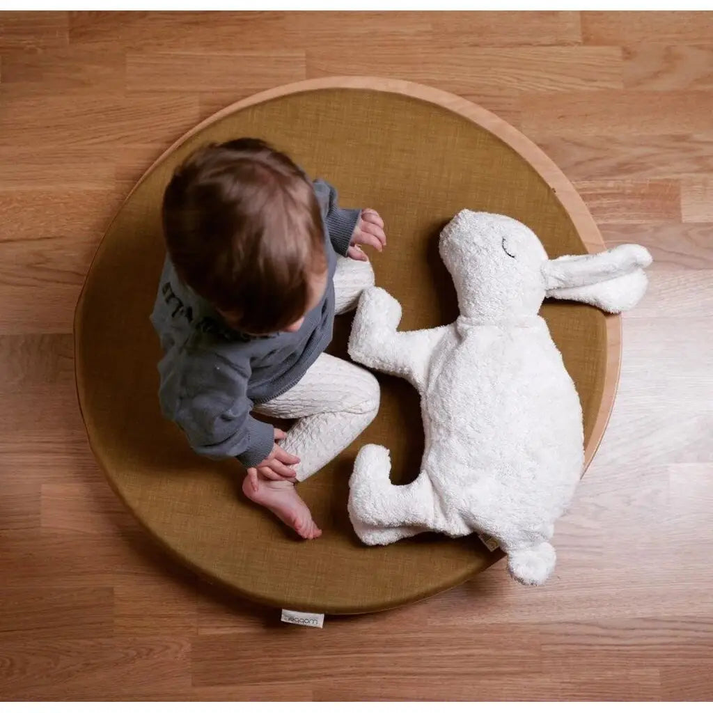 A toddler sits cross-legged on a round brown cushion beside a Senger Naturwelt Cuddly Animal - White Bunny, on a wooden floor. The photo is taken from above.