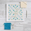 A Scrabble Bianco Edition game board set on a white surface with gold and blue grid squares, featuring letter tiles spelling “PLAY,” “OCEAN,” and “SHORE,” with four tile racks, a teal drawstring bag, a timer, and a score pad.