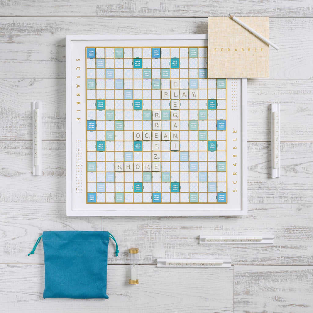 A Scrabble Bianco Edition game board set on a white surface with gold and blue grid squares, featuring letter tiles spelling “PLAY,” “OCEAN,” and “SHORE,” with four tile racks, a teal drawstring bag, a timer, and a score pad.