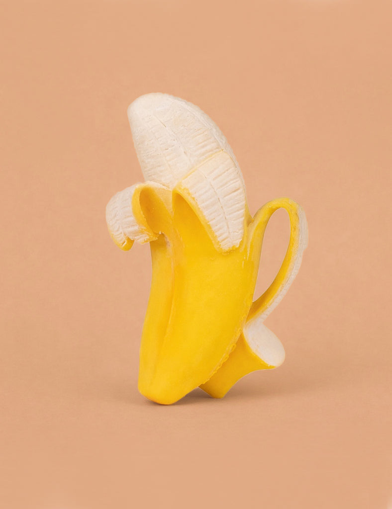 The Ana Banana Teether features a banana with its peel partially open, but the fruit is replaced by a white ceramic coffee cup on a solid light brown background, creating a whimsical sensory play toy.
