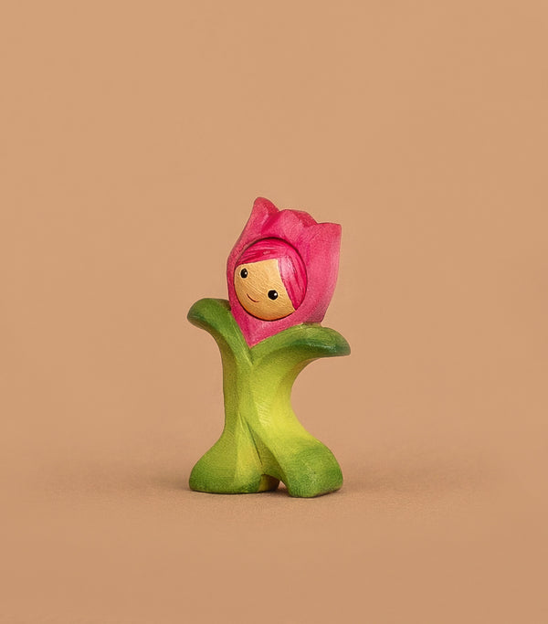 The Bumbu Handmade Wooden Flower Child Tulip, featuring a smiling face with a pink flower hat and green petal outfit, stands upright against a beige background—perfect for open-ended play. Ships in one week.