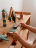 A child’s hands play with the Sabo Concept Sea Adventure Set on a shelf, featuring a lighthouse, sailboat, rocket, trees, and airplane in a softly lit and cozy scene.