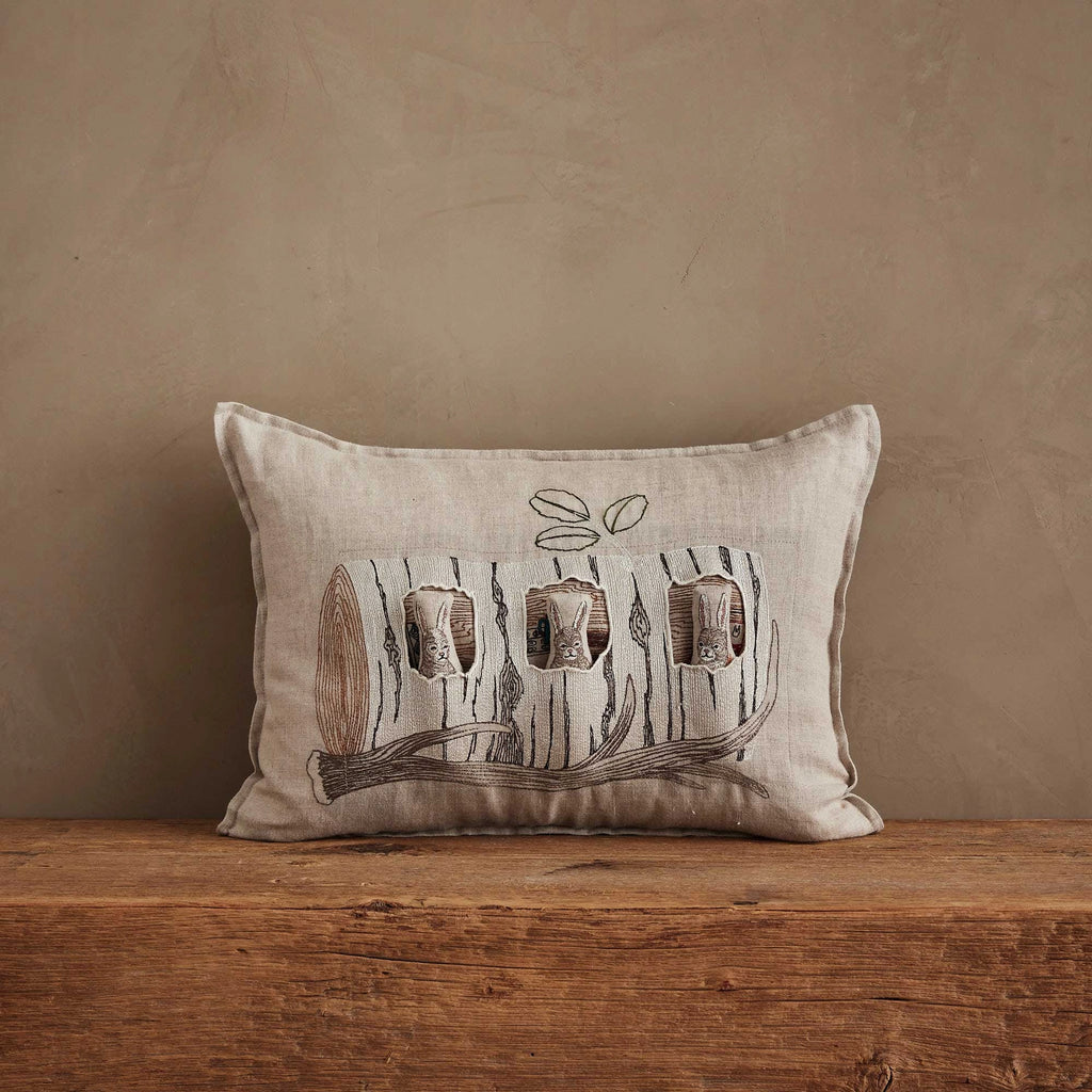 The Coral & Tusk Aspen Log Bunnies Pocket Pillow is beige with a rustic log illustration and features three removable bunny pals peeking out of holes, displayed on a wooden surface against a neutral, textured wall.