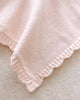 A close-up of the Cuddle + Kind Organic Cotton Baby Bunny Lovey (Blush), featuring soft, light pink hand-knit fabric with a scalloped edge and delicate ribbed border, displayed on a textured beige surface.