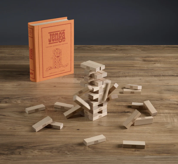 A wooden Jenga tower with some blocks removed sits on a table beside the orange, book-shaped Jenga Vintage Bookshelf Edition box—perfect for family game night.