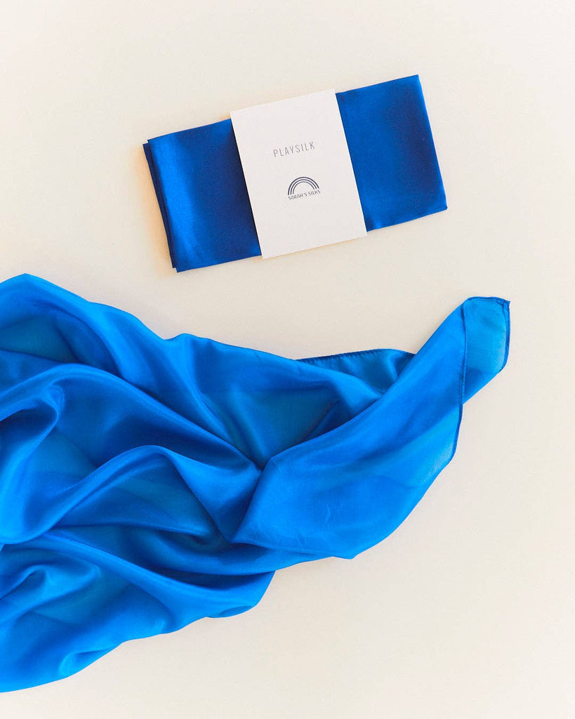 A vibrant royal blue pure silk scarf, Sarah's Silks Solid Color Playsilk - Royal, is partially folded on a light surface. Above it sits another neatly folded Playsilk wrapped with a white "PLAYSILK" label and simple logo. Ideal for open-ended play.