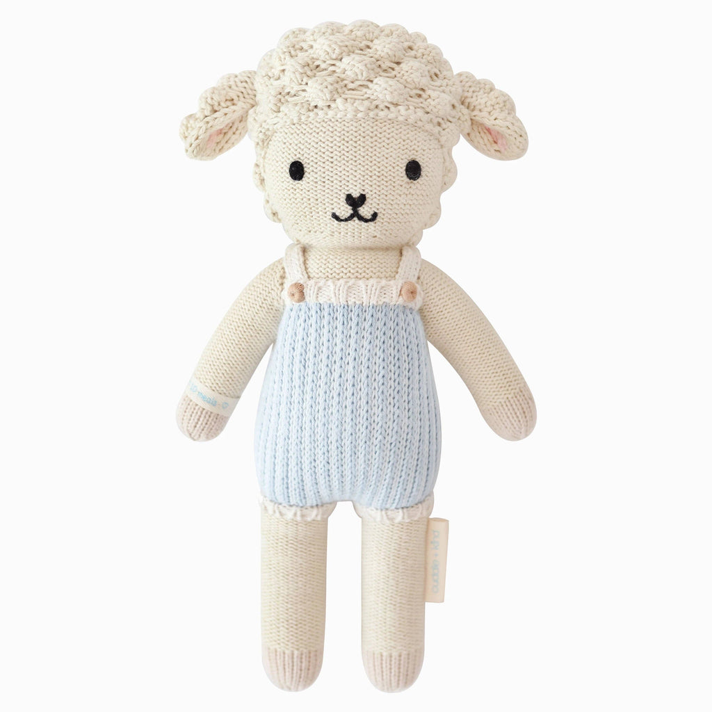 Cuddle + Kind Benjamin the Lamb is a hand-knit cotton doll with cream-colored wool, floppy ears, black embroidered features, and light blue knit overalls. This charming toy stands upright against a white background.