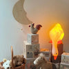 A cozy children's room features the Duck Looking Down Lamp glowing softly, toy blocks, a wicker basket of stuffed animals, a knitted horse, a fabric rooster, and a wooden moon wall decor with a sleepy face.