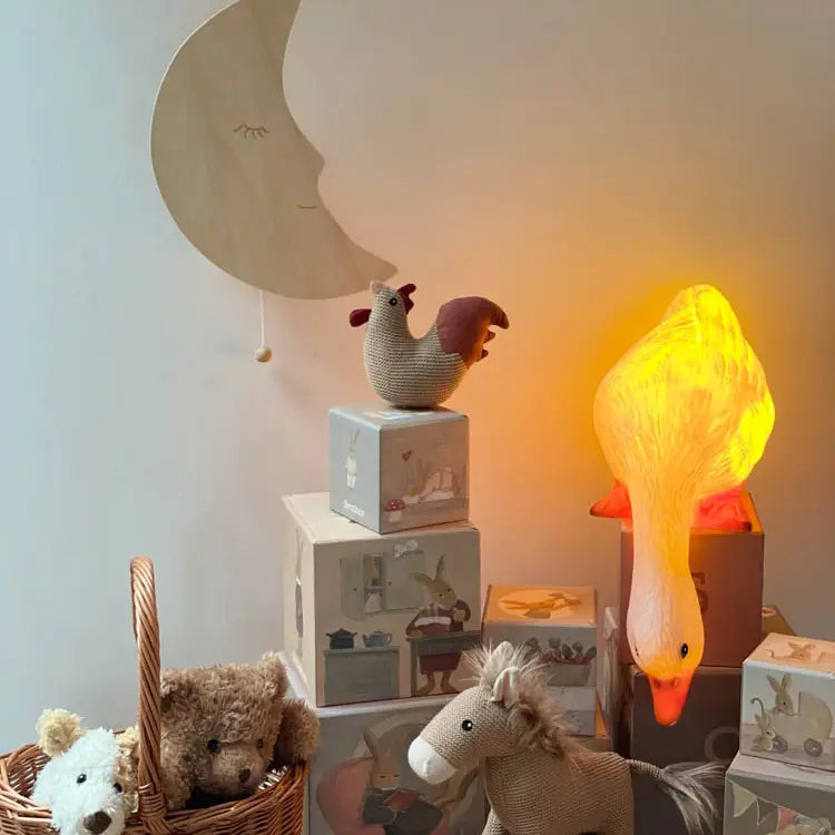 A cozy children's room features the Duck Looking Down Lamp glowing softly, toy blocks, a wicker basket of stuffed animals, a knitted horse, a fabric rooster, and a wooden moon wall decor with a sleepy face.