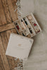 An open Sabo Concept Christmas Ornaments box rests on a wooden floor, displaying hand-painted ornaments and a nutcracker ornament set nestled in shredded paper, with part of a fringed white rug nearby.