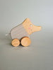 The Pull and Push Toy Little Wild Boar, handcrafted from sustainable materials, rests on a gray surface. It features a stylized animal with a curved body adorned with light brown stripes and has a smiling face, two eyes, and pink cheeks painted on it.