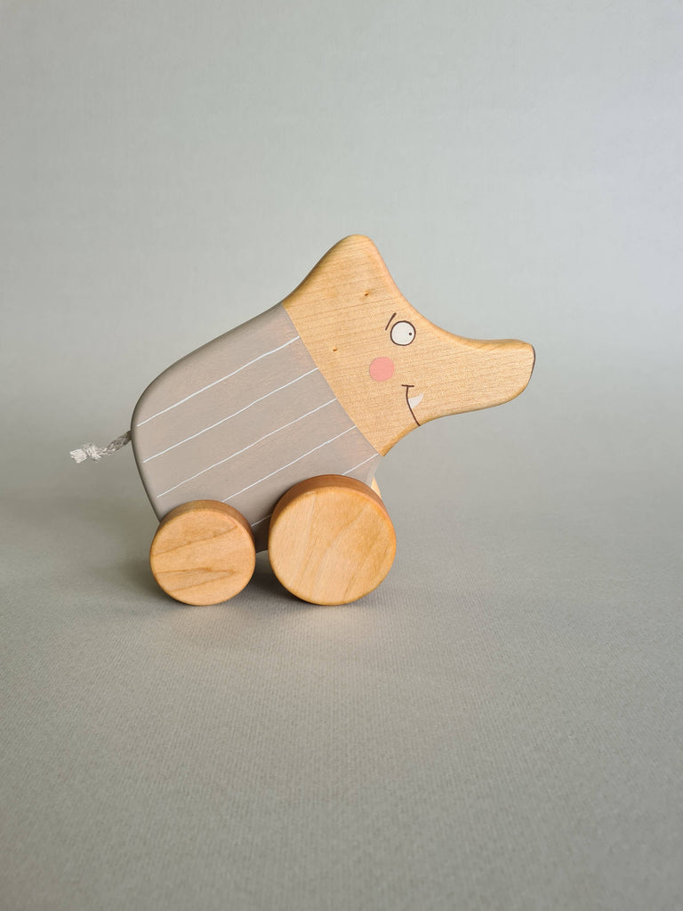 The Pull and Push Toy Little Wild Boar, handcrafted from sustainable materials, rests on a gray surface. It features a stylized animal with a curved body adorned with light brown stripes and has a smiling face, two eyes, and pink cheeks painted on it.