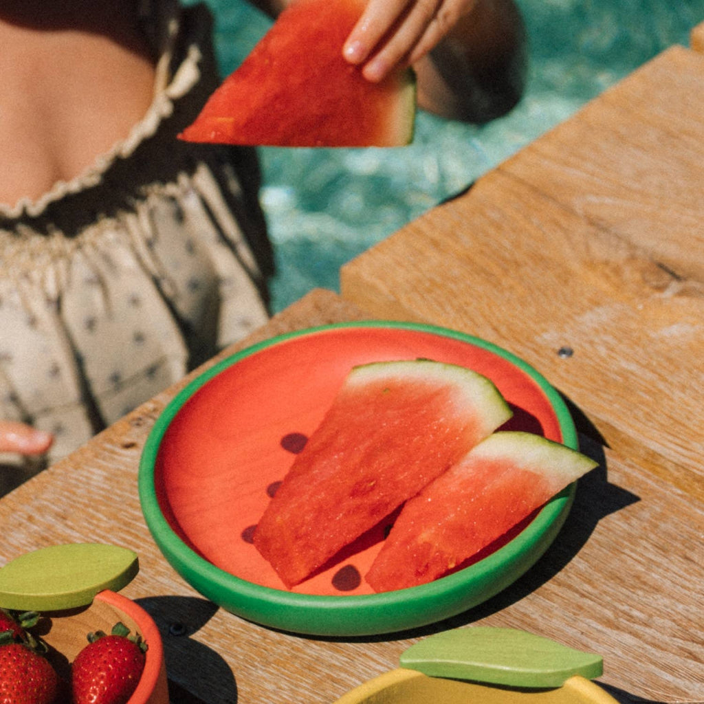 A person holds a slice of watermelon above a wooden table where the Wally the Watermelon Plate & Spoon Set, featuring two more slices, sits nearby fresh strawberries and non-toxic tableware in the background.