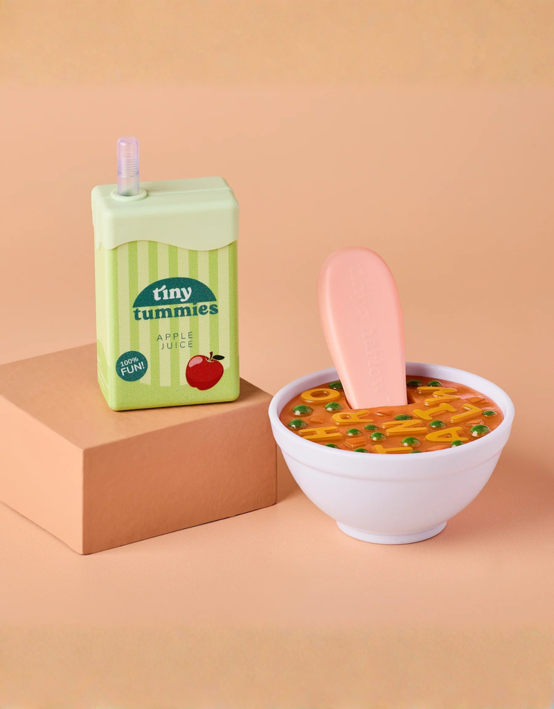 The Tiny Tummies Set - Alphabet Soup Bowl and Juice Box For Dolls, featuring a toy apple juice box and a bowl of toy alphabet soup with a large pink spoon, is displayed on a peach-colored surface, with the juice box positioned atop a cube-shaped platform.