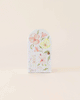 A small, arched white door featuring large pastel flowers, a brass knob, and an illustration of Tiny Emma the Mouse with "emma the mouse" text, handmade in Peru. Set against a plain beige backdrop. Product: Cuddle + Kind Tiny Emma the Mouse.