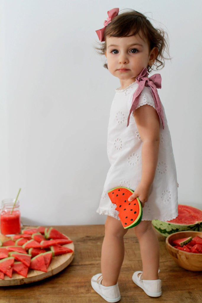 A young child in a white dress with pink bows holds Wally the Watermelon Teether. Behind her are plates of fresh watermelon slices and a glass of watermelon juice on a wooden surface.