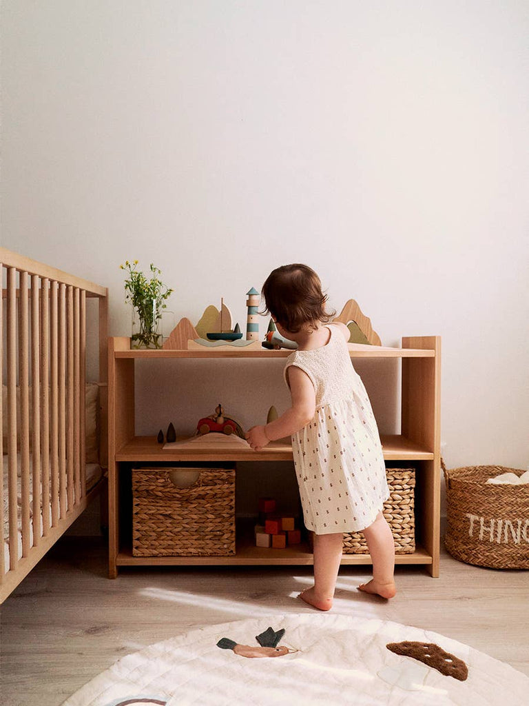 A toddler in a white dress stands near a wooden shelf with toys, baskets, and the Sabo Concept First Journey play set, inviting imagination and open-ended play in a cozy nursery beside a crib.