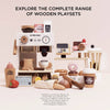 The Barista Café Shop wooden playset, featuring a coffee machine, blender, cash register, play food, and drinks, is displayed on a light background. Text above: "Explore the complete range of wooden playsets." Ships in one week.
