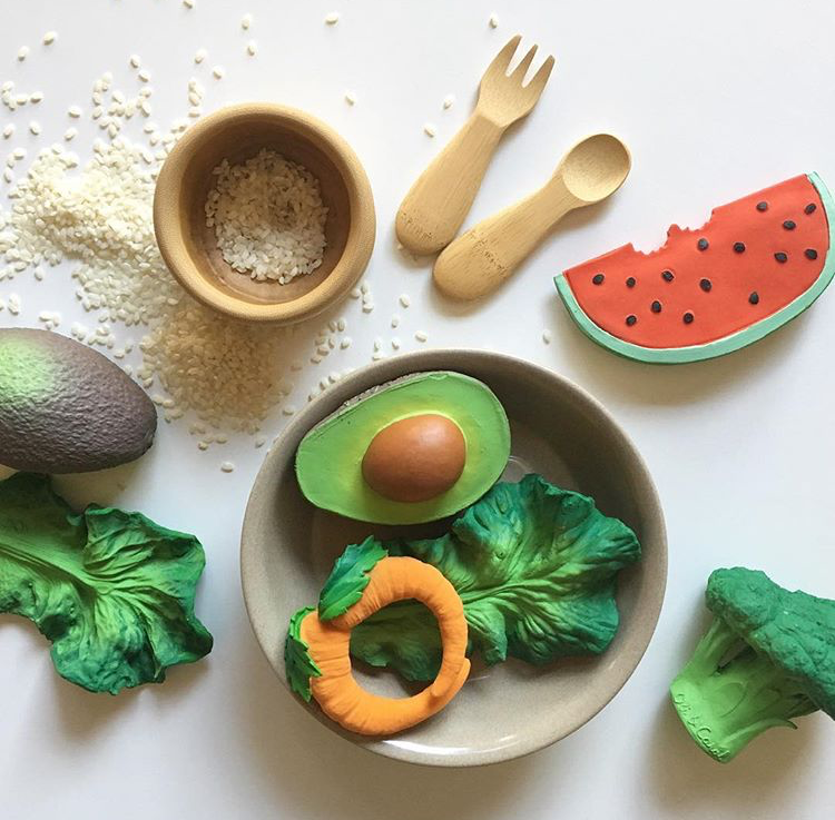 A flat lay featuring Arnold the Avocado Teething Toy among toy fruits, vegetables, rice, a wooden bowl, and bamboo cutlery on a white surface—perfectly arranged for sensory play.