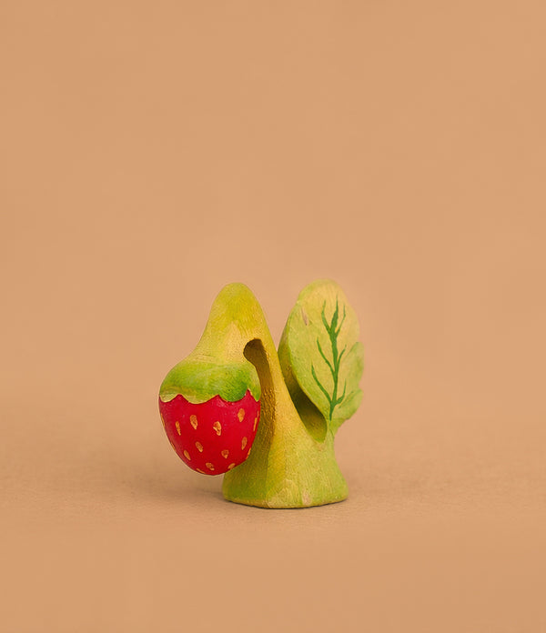 The Bumbu Handmade Wooden Strawberry Plant, inspired by Montessori education, features a green leaf and red strawberry with yellow seeds, set against a plain light brown background. Ships in one week.
