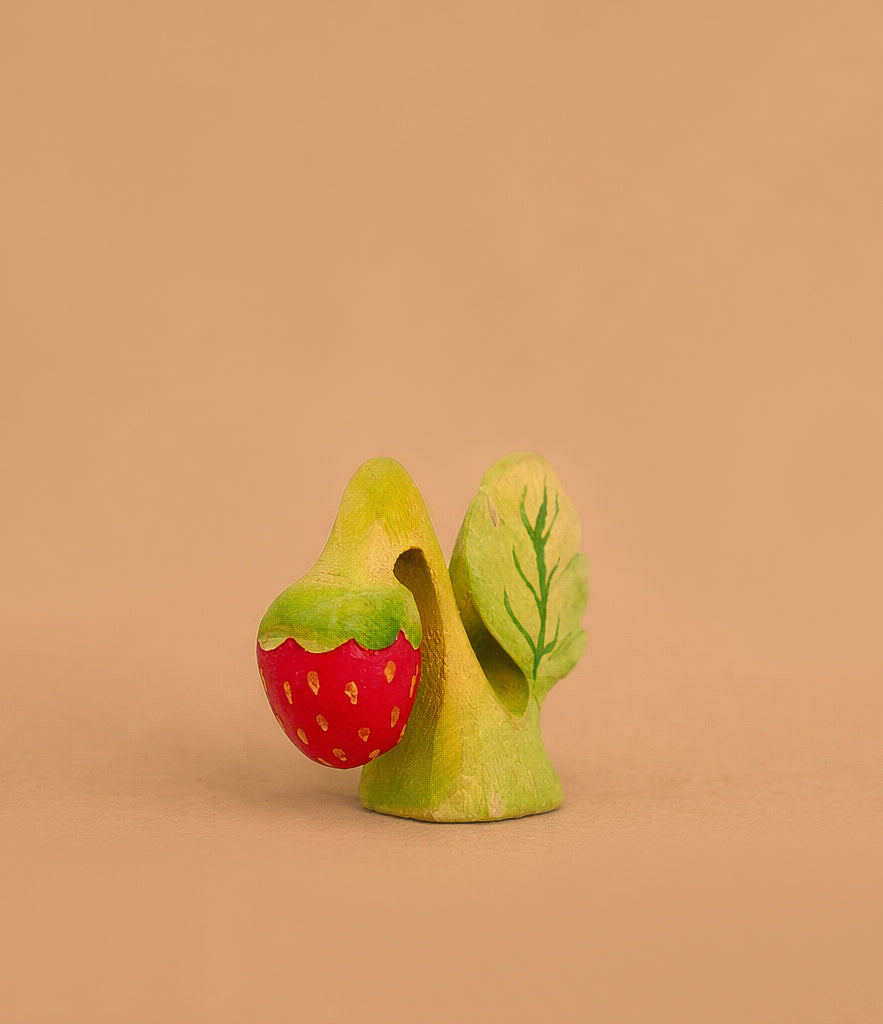 The Bumbu Handmade Wooden Strawberry Plant, inspired by Montessori education, features a green leaf and red strawberry with yellow seeds, set against a plain light brown background. Ships in one week.