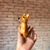 A hand holds a small, handmade Shiba-Inu Dog Finger Puppet made of wool felt, in front of a white brick wall background.