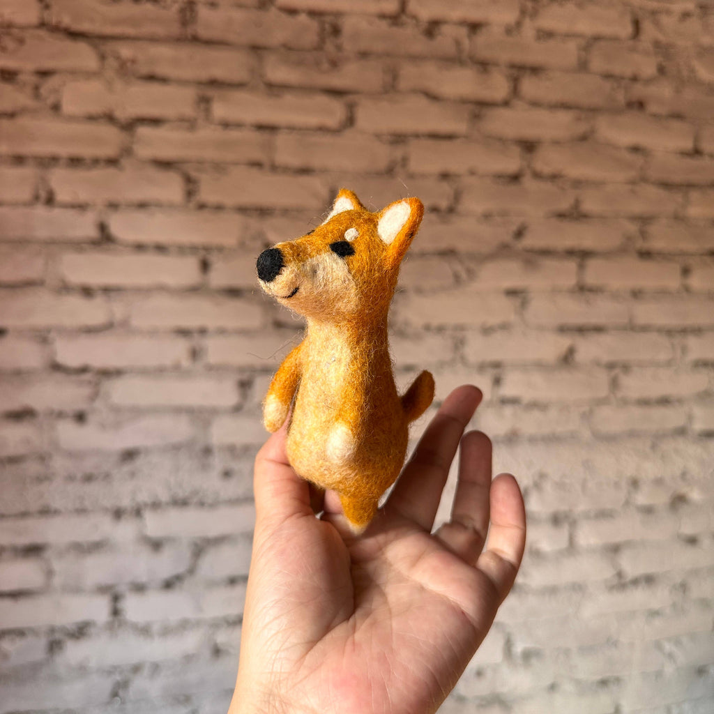 A hand holds a small, handmade Shiba-Inu Dog Finger Puppet made of wool felt, in front of a white brick wall background.