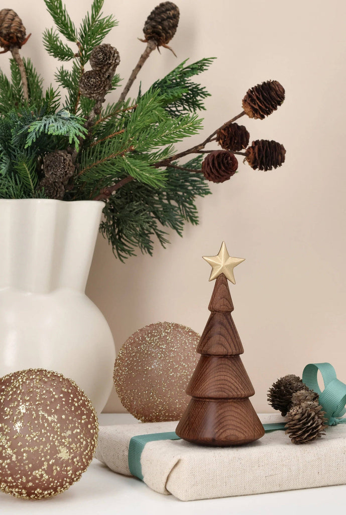 Spring Copenhagen The Christmas Tree, Thermo Beech, a small tiered wooden tree topped with a gold star, sits on a gift box, surrounded by two glittery gold ornaments and pine branches in a white vase.