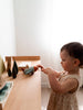 A young child in a beige dress plays with a wooden toy airplane and the Sabo Concept Aviator Mouse at a wooden table, surrounded by wooden trees in a softly lit room.