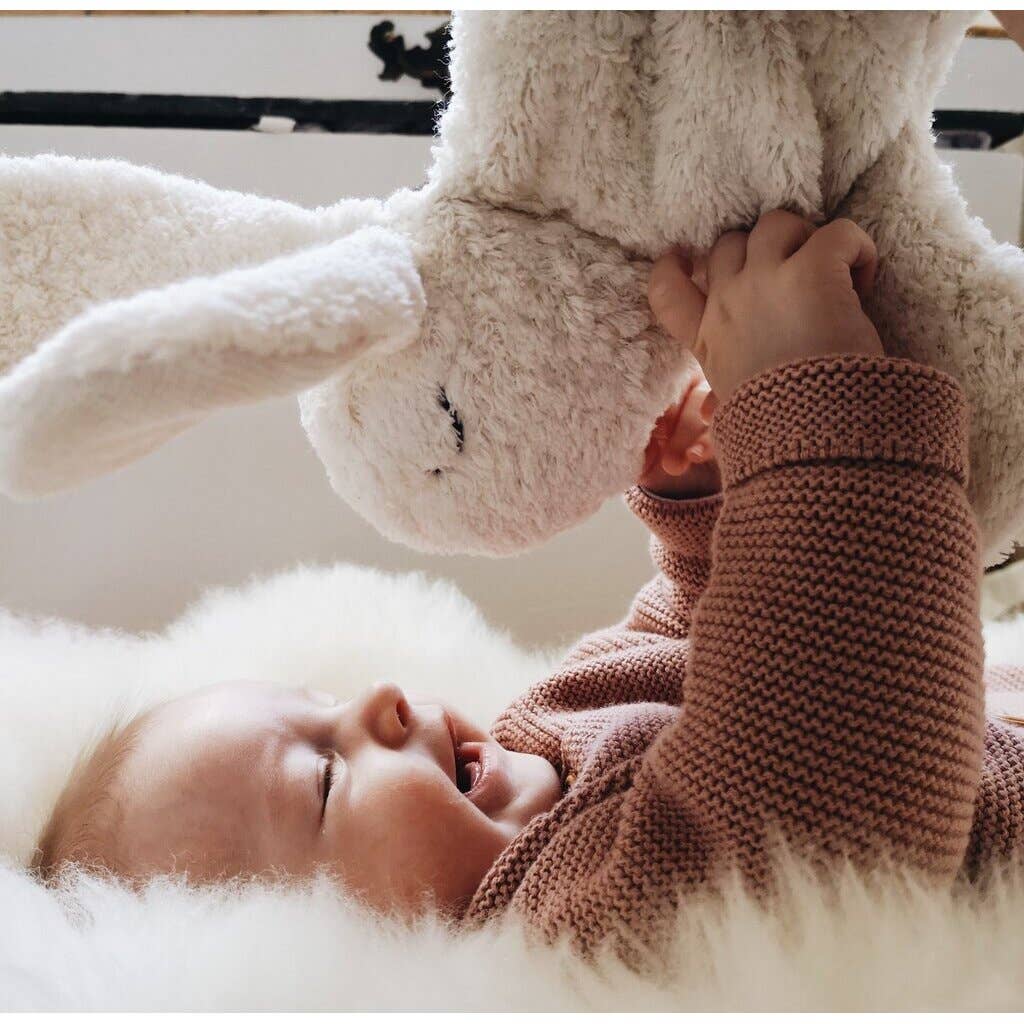 A smiling baby in a pink knitted sweater lies on a soft, fluffy surface, playing with a large Senger Naturwelt Cuddly Animal - White Bunny.