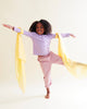 A young child with curly hair balances on one leg, arms outstretched, smiling as colorful Sarah's Silks Solid Color Playsilk - Sunshine is tied to each arm—perfect for open-ended play against a plain light background.