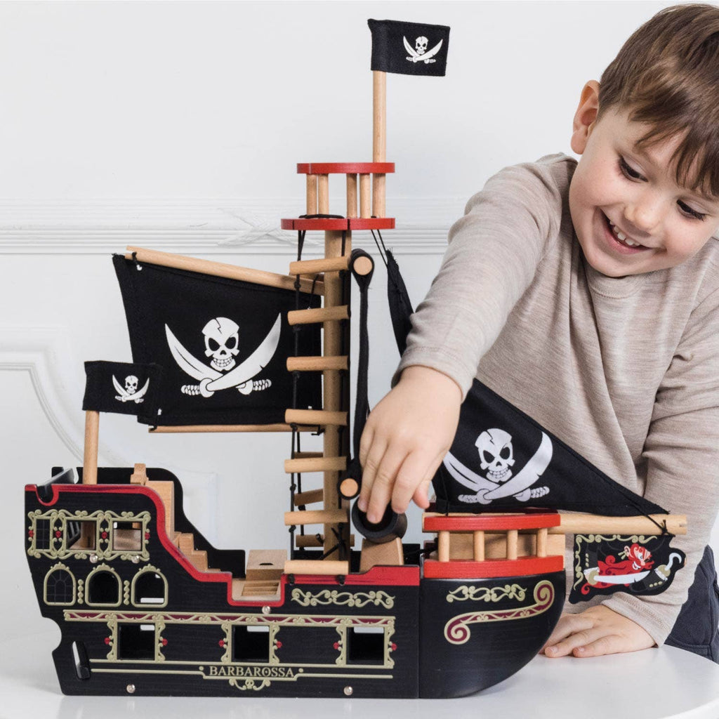 A smiling young boy plays with the Barbarossa Pirate Ship & Characters, a wooden pirate ship toy adorned with skull flags, gold accents, and red trim, inside a bright room. Ships in one week.