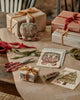 A wooden table with wrapped gifts tied with velvet ribbons, greeting cards, red scissors, spools of ribbon, and the Coral & Tusk Mouse with Wreath Ornament sets a cozy and festive holiday gift-wrapping scene.