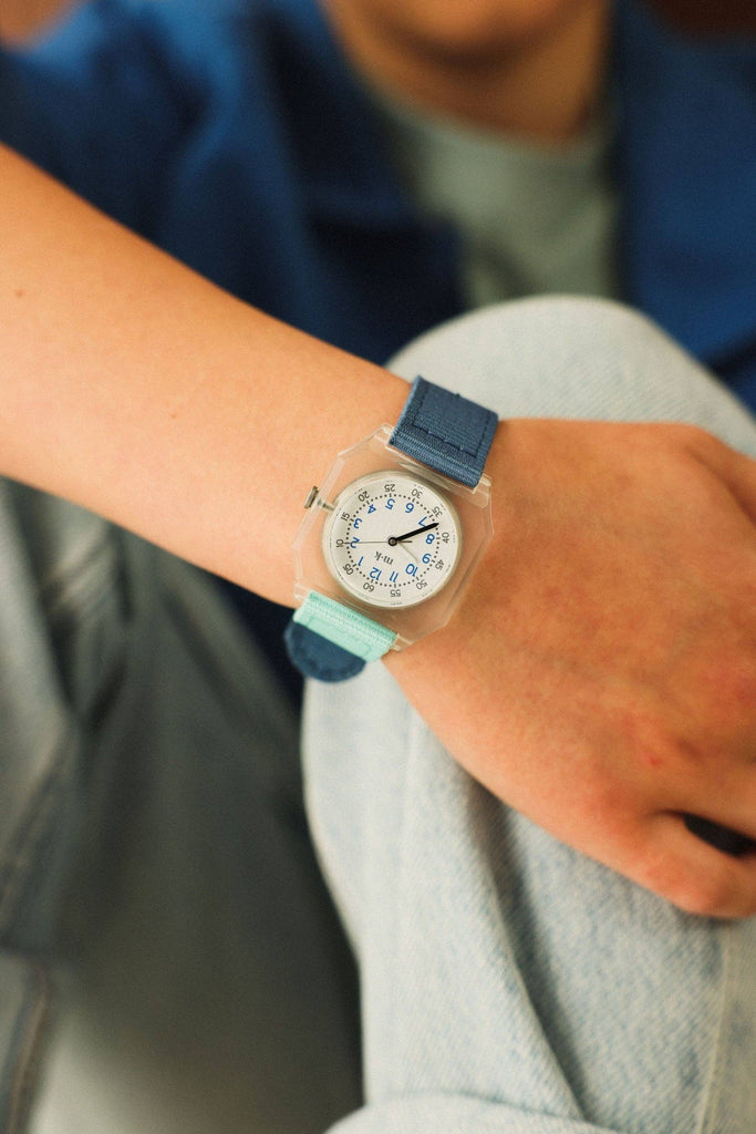 A person in light blue jeans and a blue jacket displays their wrist wearing the Mini Kyomo Watch - Blue Ocean, which features a blue and turquoise strap and a white dial with black markings.