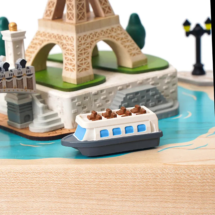 The Paris Music Box features a small toy boat floating on blue, with miniature wooden models of the Eiffel Tower and Paris landmarks, creating a charming, classic scene inspired by hand-cranked music boxes.